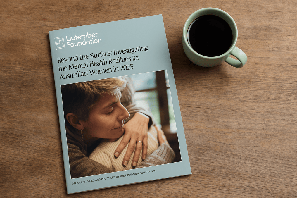 A printed research report by the Liptember Foundation titled "Beyond the Surface: Investigating the Mental Health Realities for Australian Women in 2025" sits on a wooden table next to a green cup filled with black coffee. The report cover features a photo of a woman embracing another person in a comforting moment.